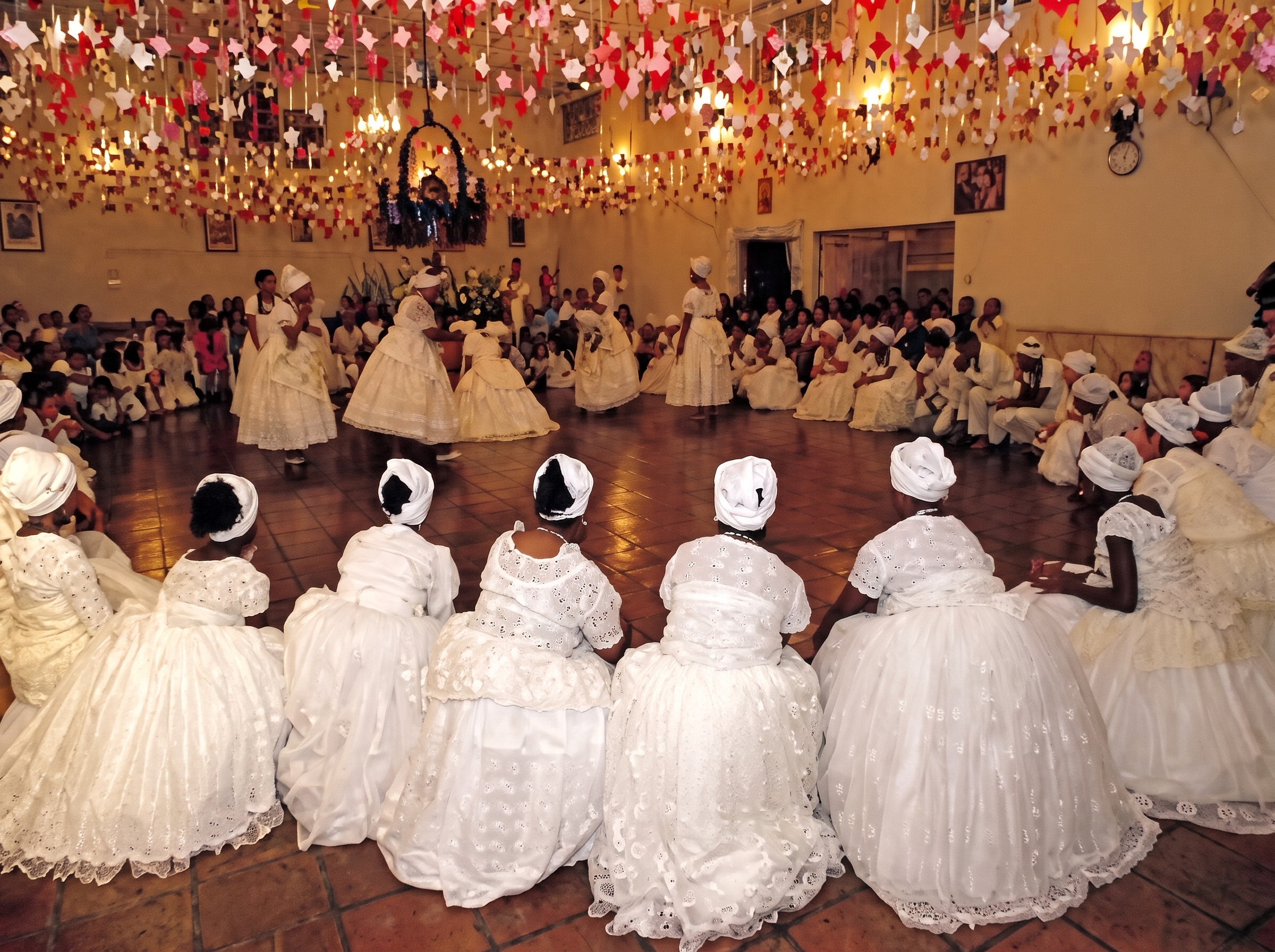 Roda de filhas de santo de branco num salão enfeitado de bandeirolas, em festa de Mina